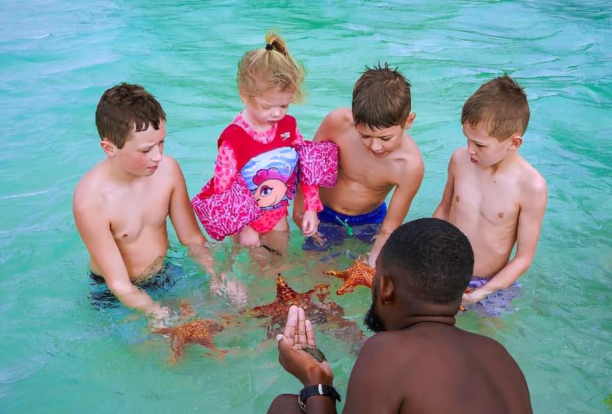 At a Starfish Point tour in Grand Cayman, children gathered around a guide holding starfish in shallow turquoise water.