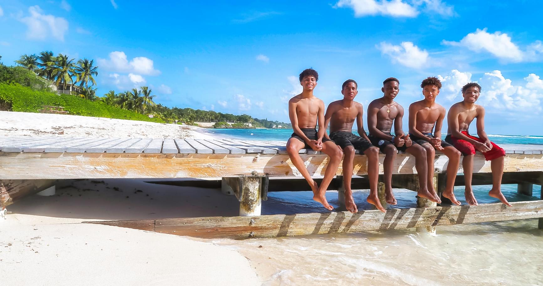 Five boys sitting on a wooden dock, enjoying a sunny day at the beach, with water and sand in the background.