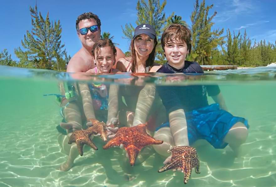 Family exploring starfish in crystal-clear waters on a sunny day at Grand Cayman Islands Starfish Point.